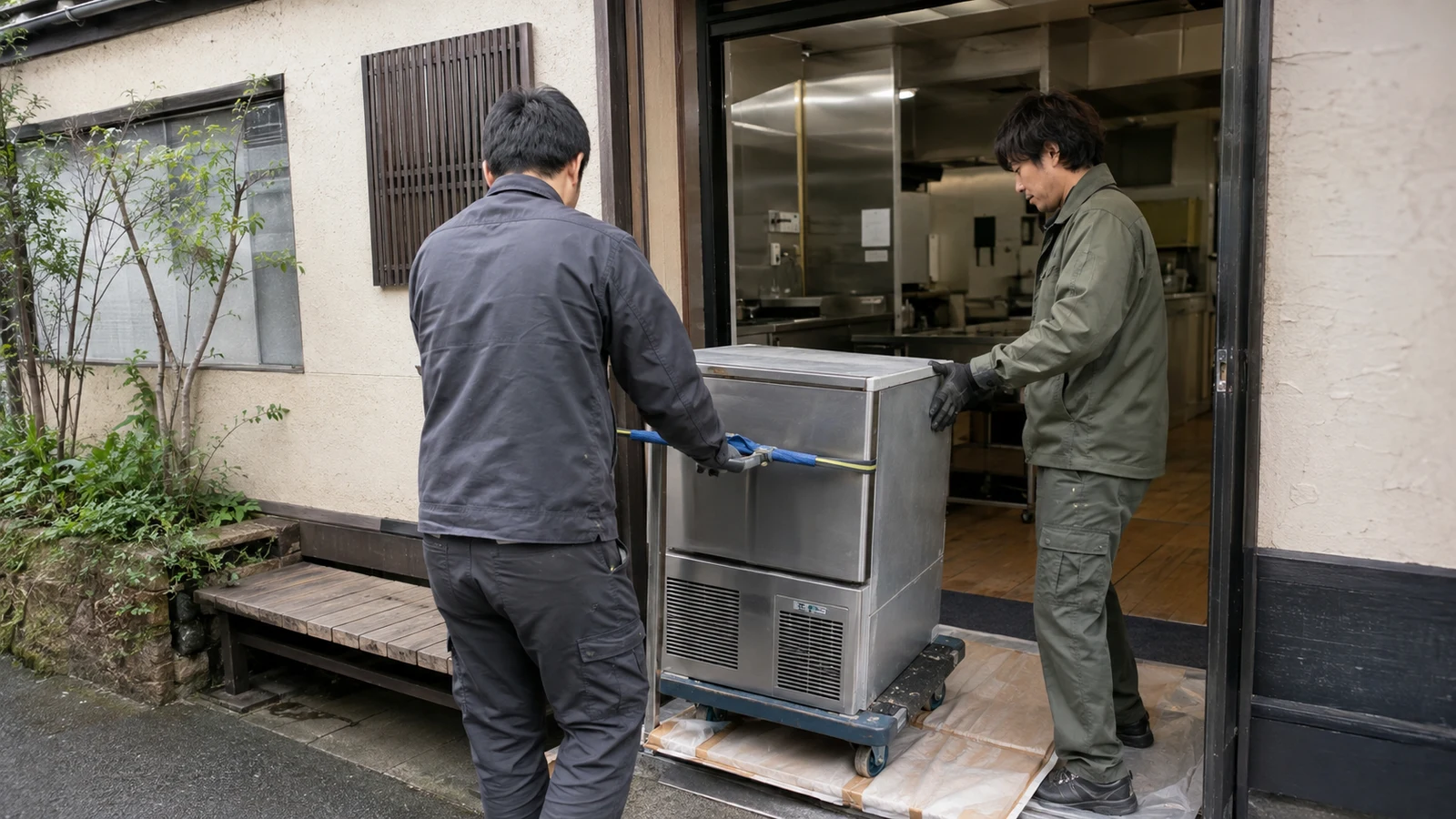 空になった飲食店の入口で、台車に載せた厨房機器を搬出する専門作業員。
