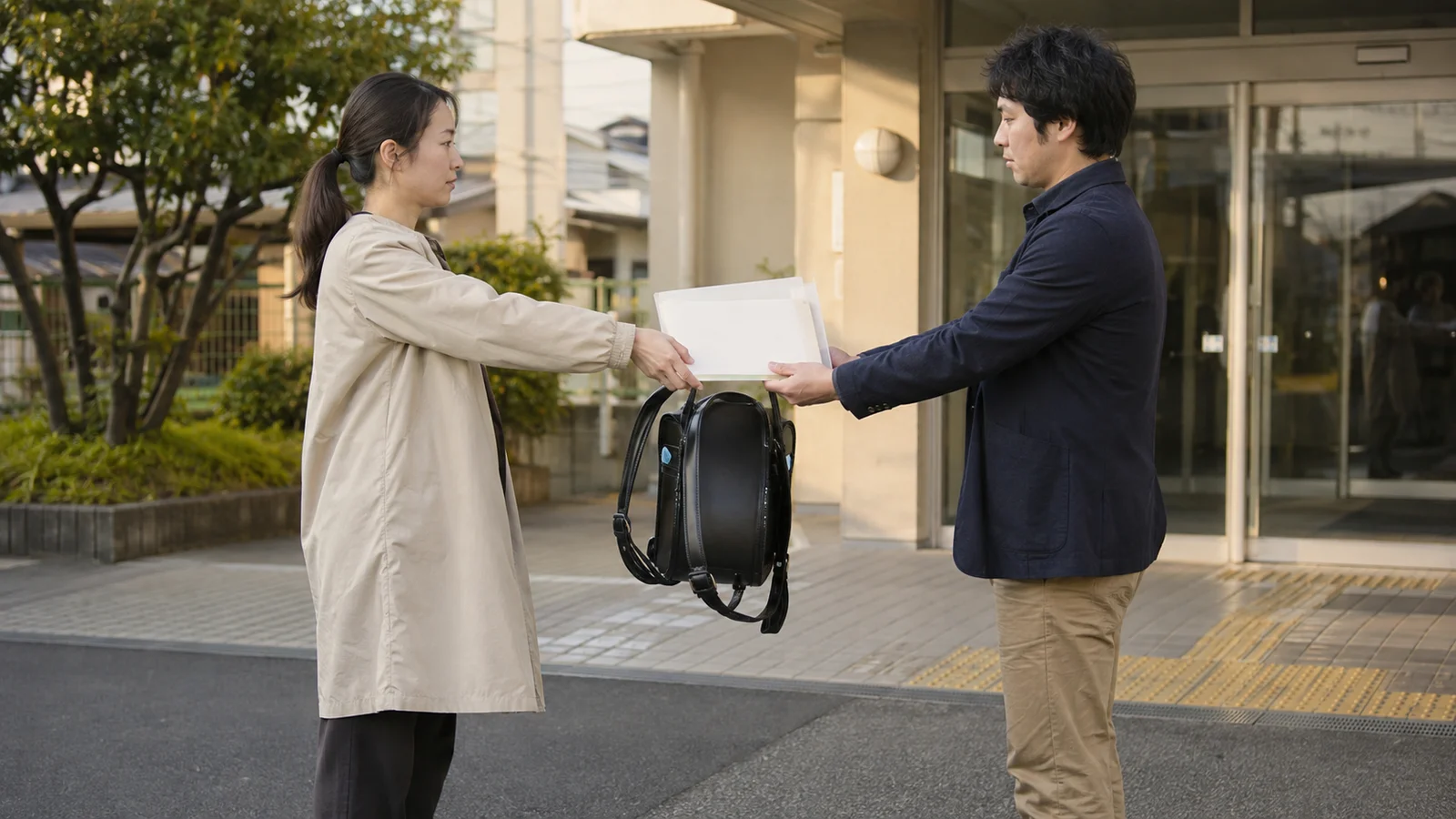 別居する父母が地域施設の前で子どもの通学鞄と予定ファイルを落ち着いて受け渡している。
