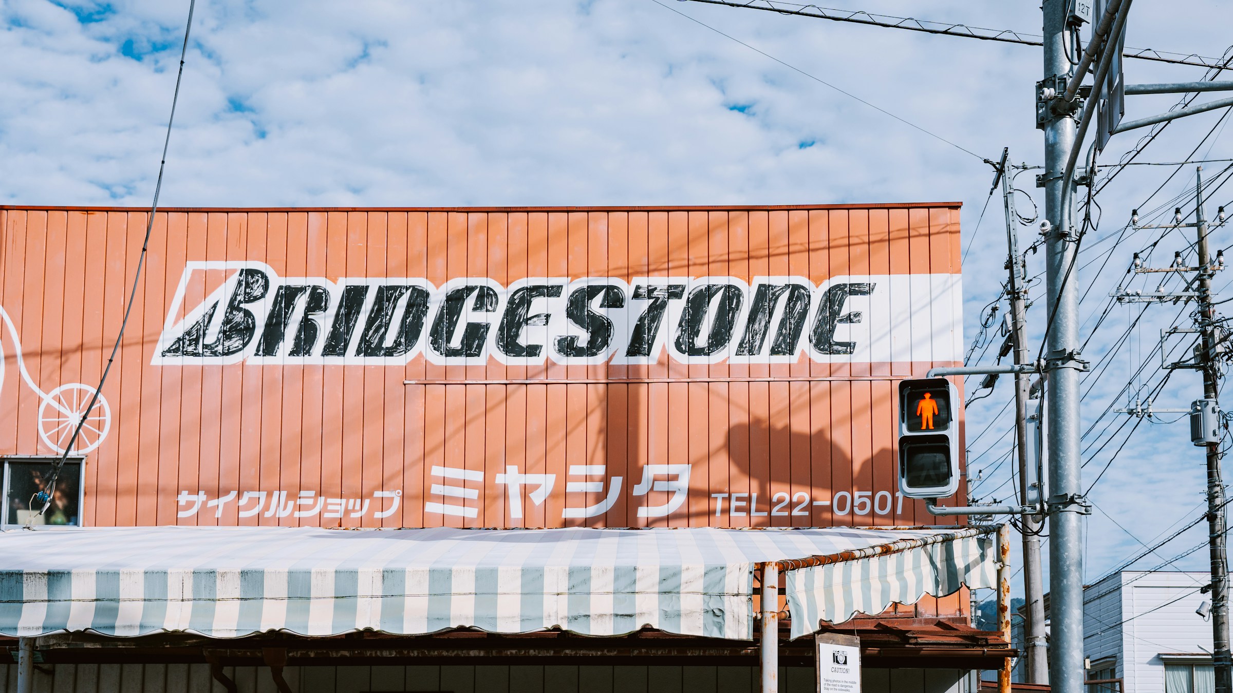 日本の地方の街の自転車店の店構え。オレンジの壁面に「BRIDGESTONE」のロゴと「サイクルショップ ミヤシタ」の店名、電話番号。赤信号と電柱が前景に映る、昭和の名残のある通り沿いの一場面。