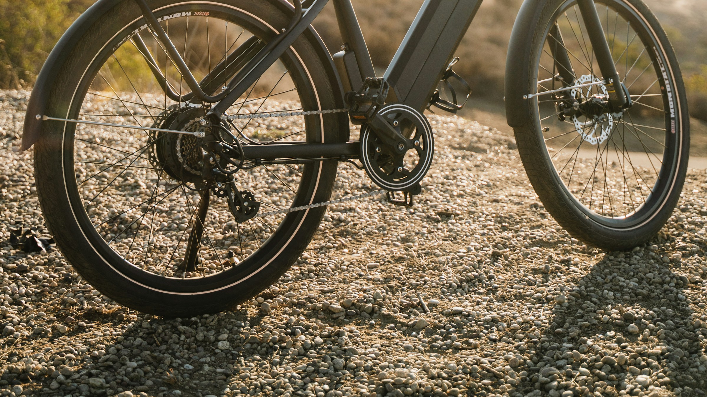 電動アシスト自転車(e-bike)の足元クローズアップ。砂利の道に止まる頑強なタイヤとミッドドライブモーター、太いフレーム、ディスクブレーキ。電動アシスト普及を象徴する一枚。