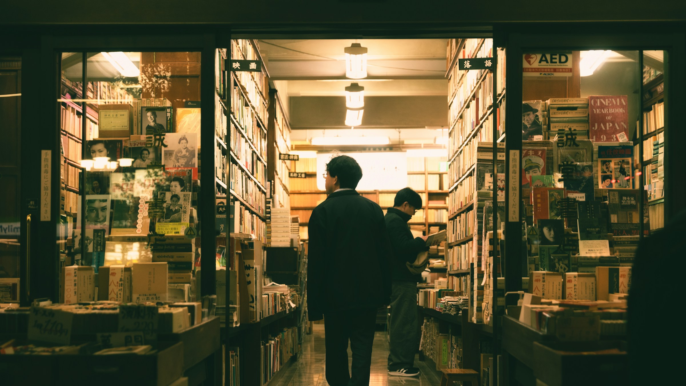 日本の書店内、薄暗い照明の下で本棚を見て本を選ぶ人々の後ろ姿。「CINEMA YEARBOOK JAPAN」の表示。