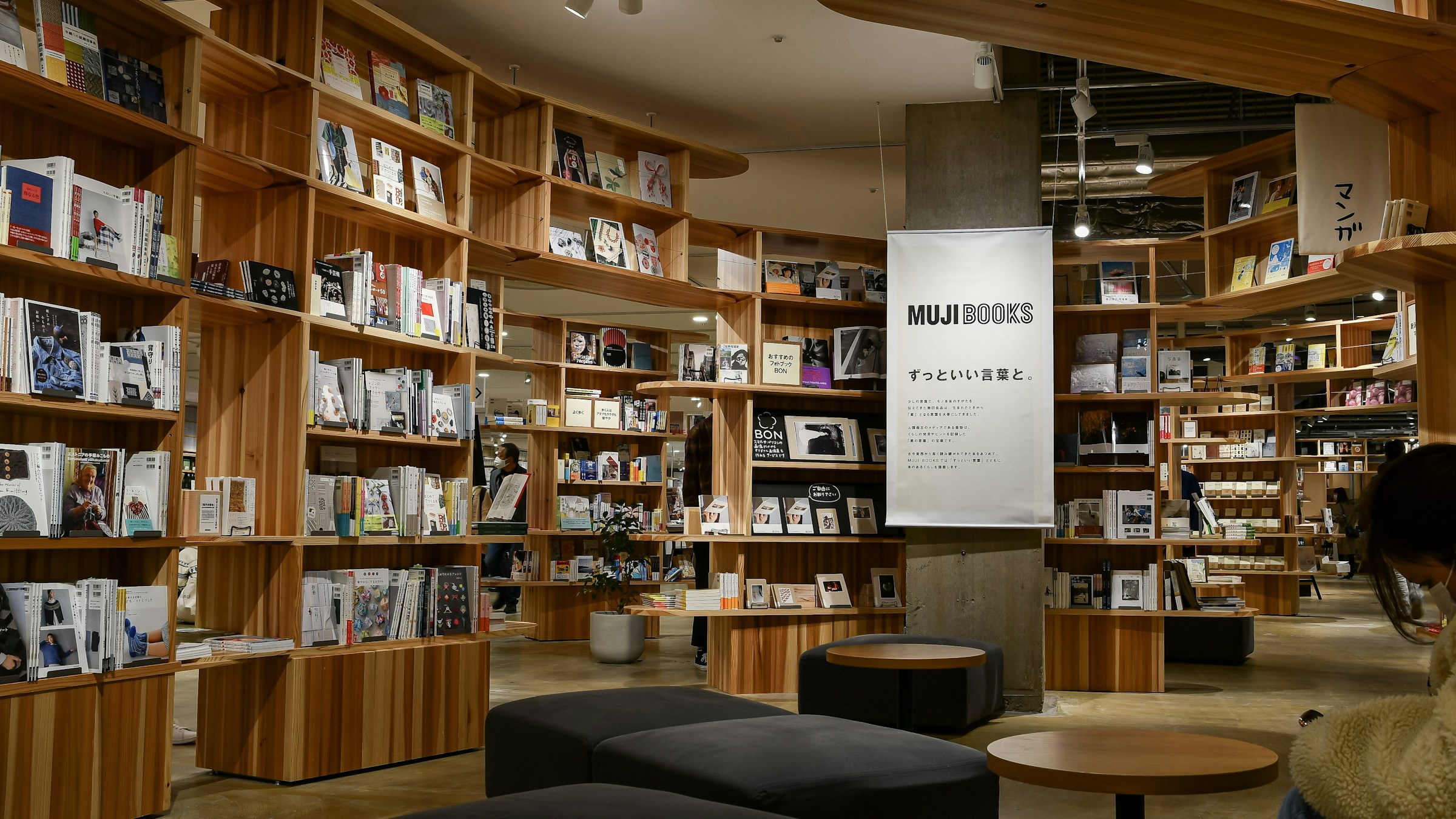 「MUJI BOOKS」店内の現代的な読書空間。木製の棚が並び、ベンチで座って本を読める設計。「ずっといい言葉と。」のキャッチコピー。