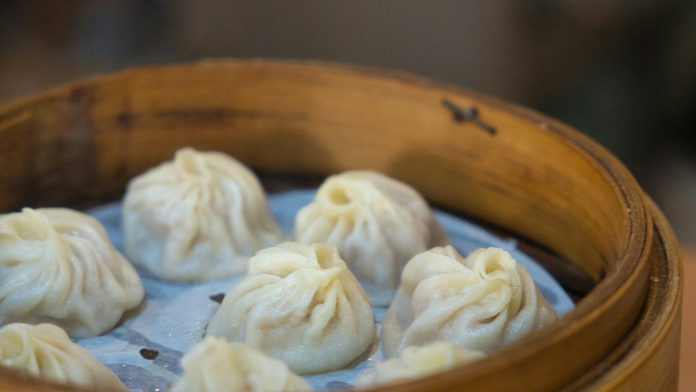 竹せいろの中に小籠包(Xiao Long Bao)が7〜8個並ぶ正面接写。竹の編み目の質感・小籠包の繊細な襞・薄い皮の透け感が鮮明、背景は深いボケで判別不可、文字・ロゴ・人物・装飾的中華街景観・赤いランタンは一切なし、6業態タイプが中華の地方料理多様性(点心専門・本格中華・台湾料理)を分け合いながら並走する構造を象徴する抽象構図。