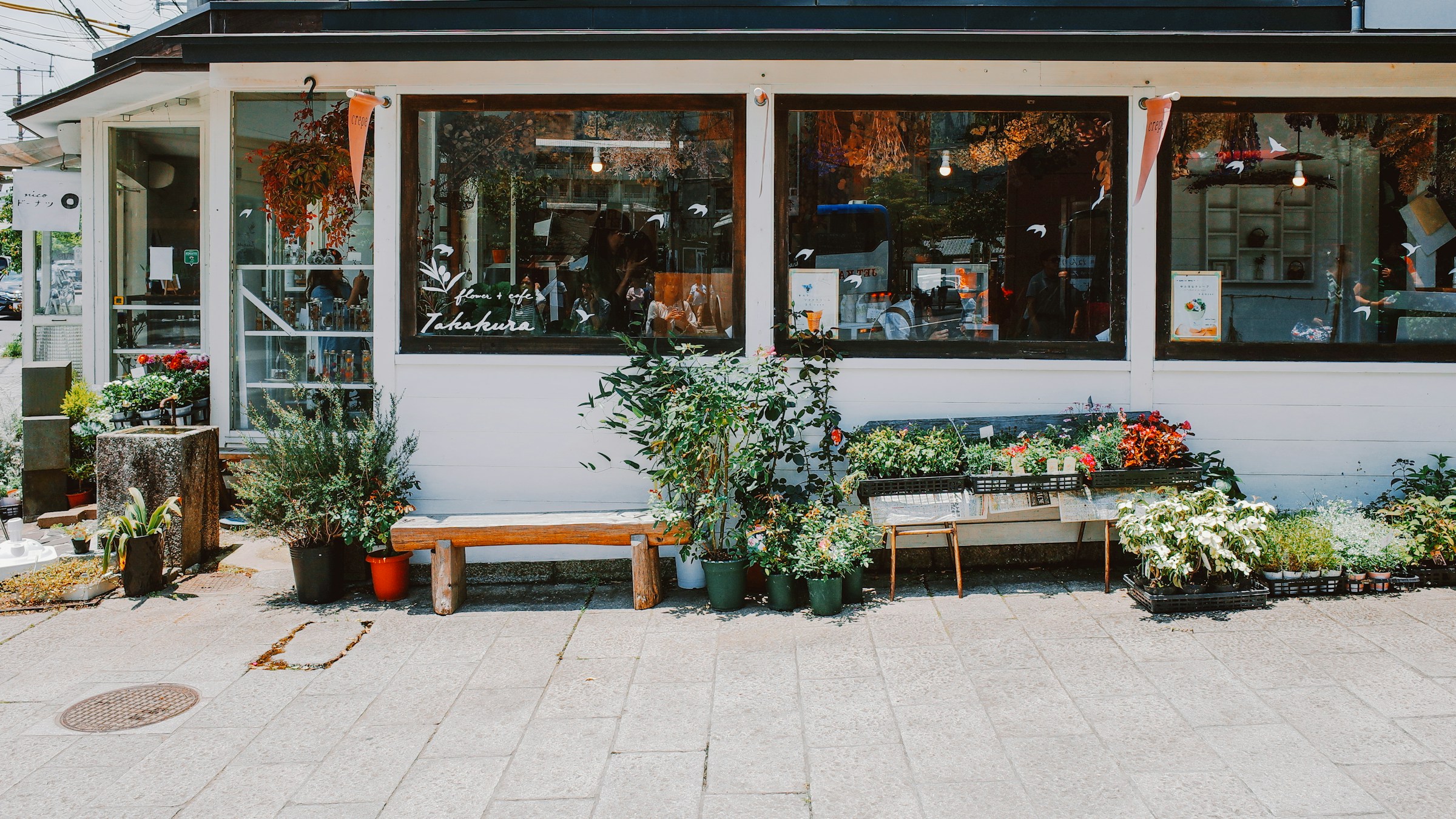 日本の地方の花屋カフェの店先。ガラス張りのファサードの前にベンチが置かれ、植木鉢やプランターが並ぶ。穏やかな日差しの中、花や緑が暮らしの導線に自然に溶け込んでいる。