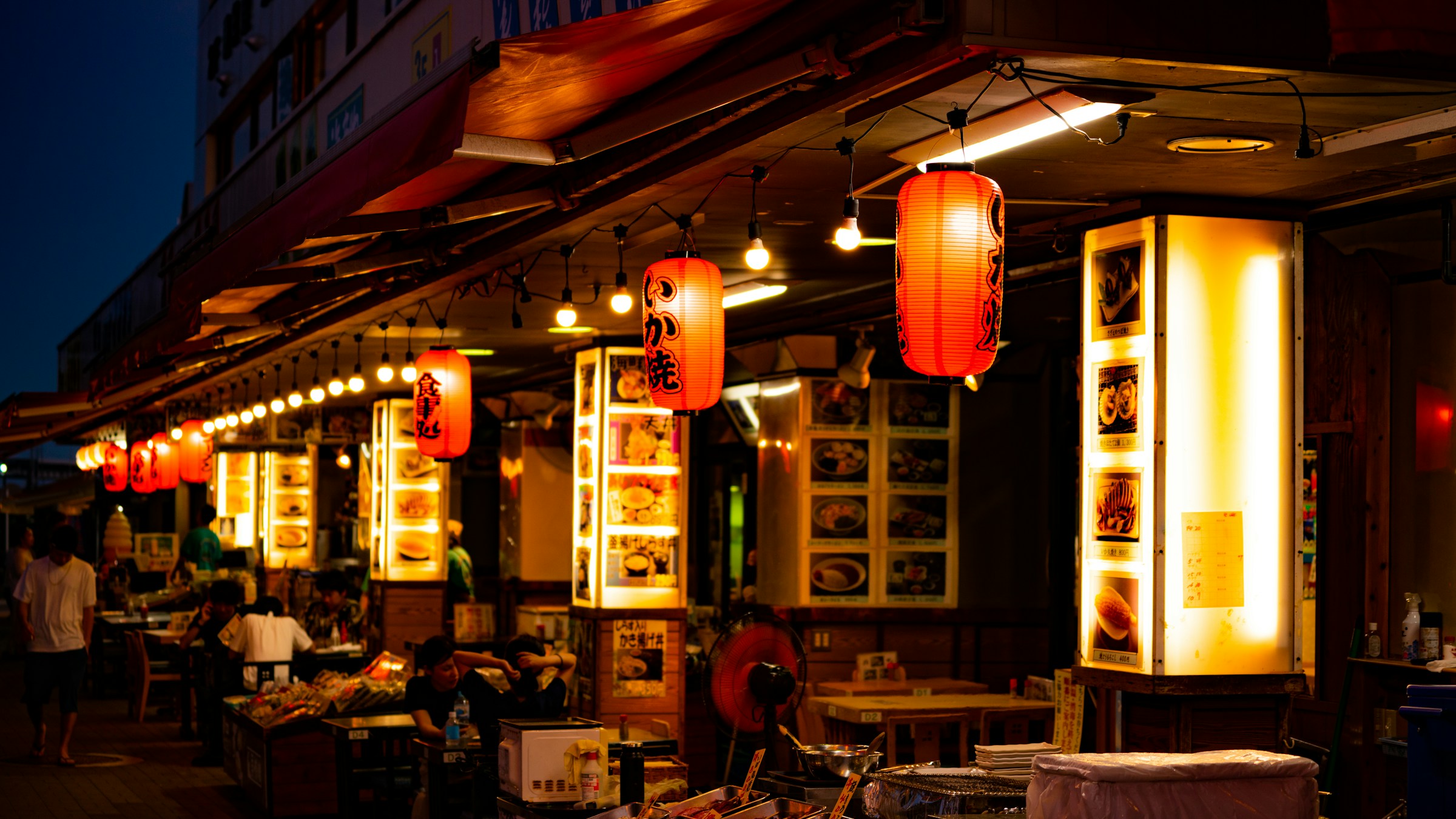 夕暮れ時の江ノ島の居酒屋通りに並ぶ赤提灯と店頭メニュー。現代の居酒屋業界の街角の風景を象徴する一枚。