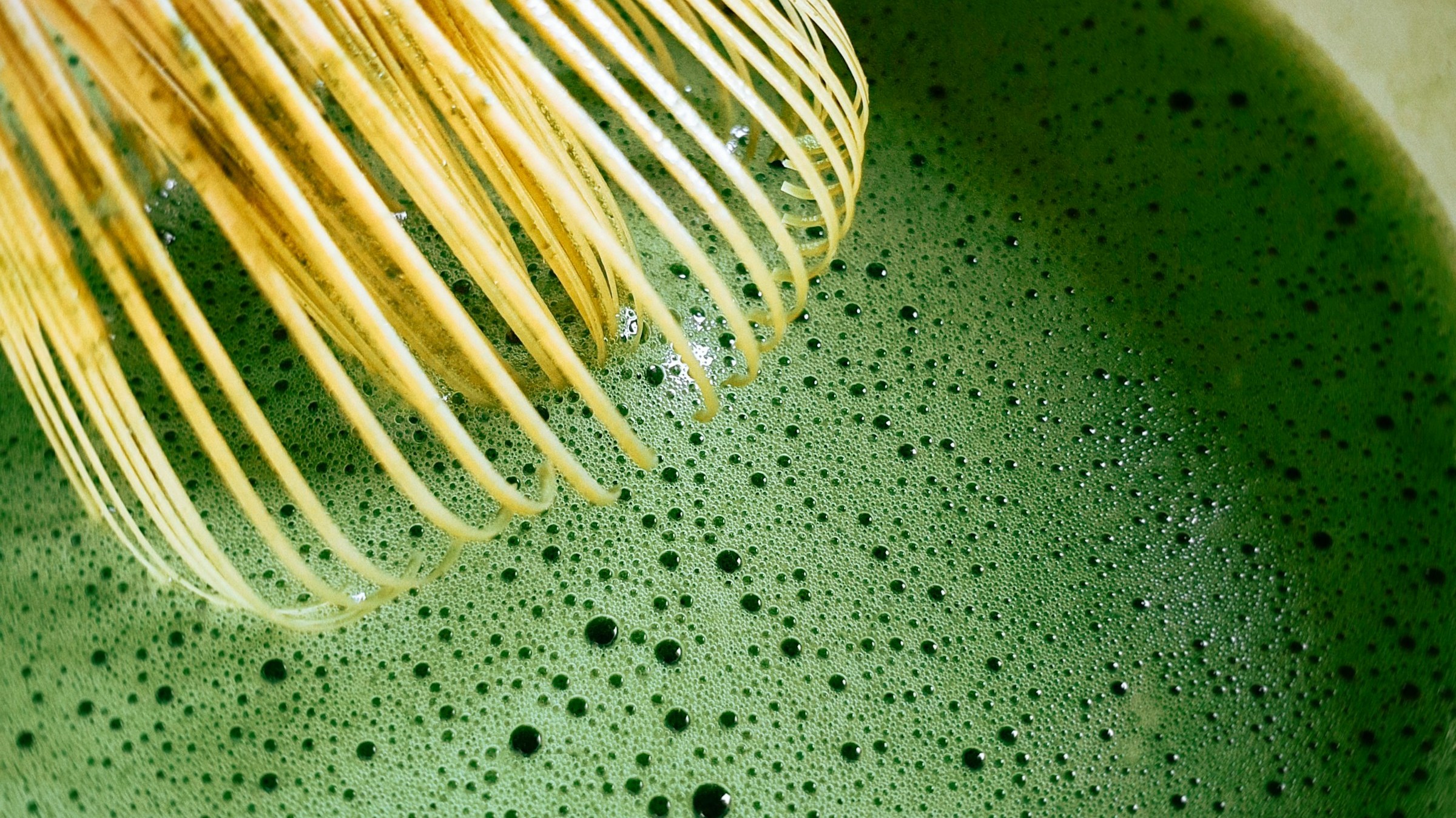 竹茶筅(chasen)の穂先と泡立つ抹茶液の極接マクロ。細やかな気泡が浮かぶ濃緑色の液面と竹細毛のディテールが浮かぶ、茶道文化の繊細な手仕事を象徴する一枚。