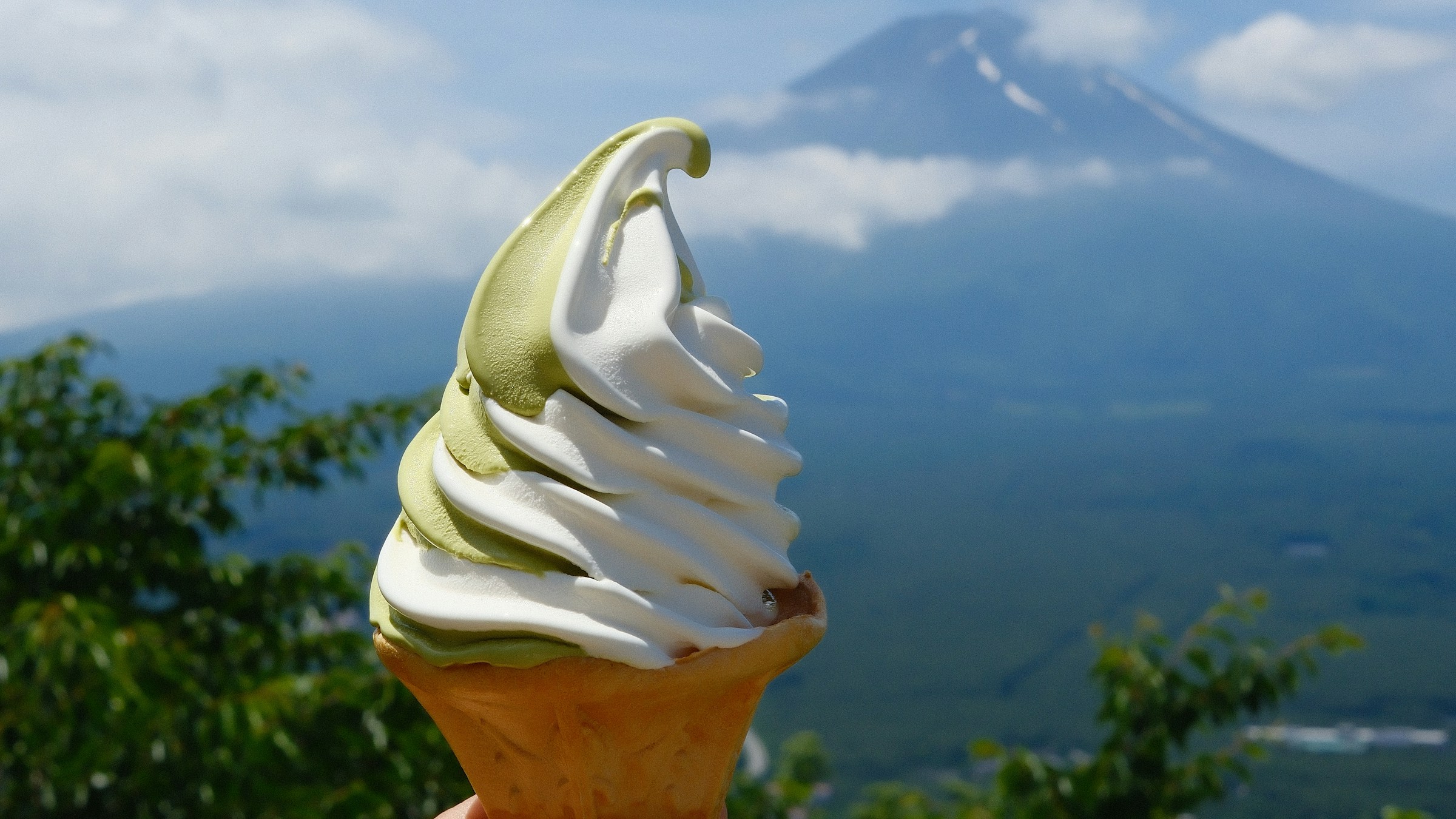 ワッフルコーンに盛られた抹茶とバニラのミックスソフトクリーム。背景に雲をまとった富士山が広がる、ご当地ソフトの一枚。