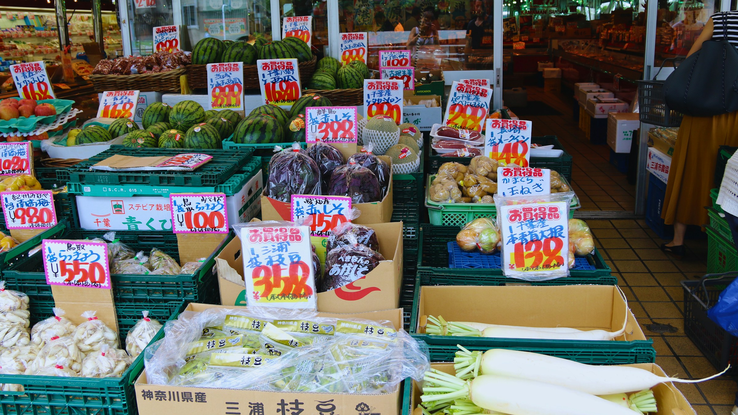 日本の農産物直売所の店頭。スイカ、なす、大根、ねぎ、ピーマンなどがプラスチックの平台に並び、「神奈川県産 三浦枝豆」「お買得品」「¥100」「¥298」「¥480」など朱書きや黄色のPOPで価格が表示されている。