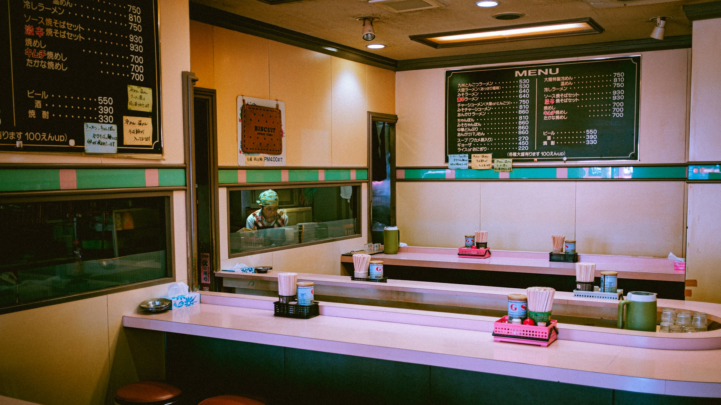 国内のラーメン店のカウンター席風景。黒板のメニューと、ピンクのカウンター、丼や調味料が整然と並ぶ、昔ながらの街のラーメン店の店内を象徴する一枚。