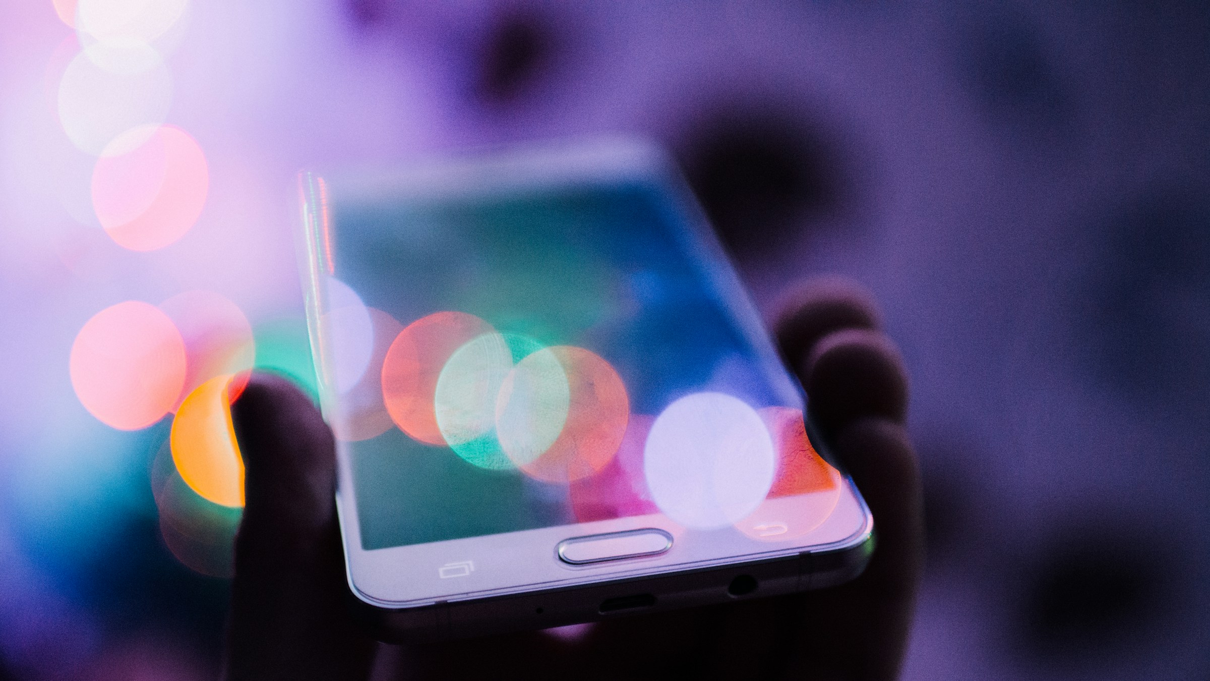 夜の街灯やイルミネーションの光が背景にぼけて広がる中、白いスマートフォンが手のひらに乗せられて画面を上に向けている。スクリーンは黒く反射し、色とりどりの光の玉が映り込む。