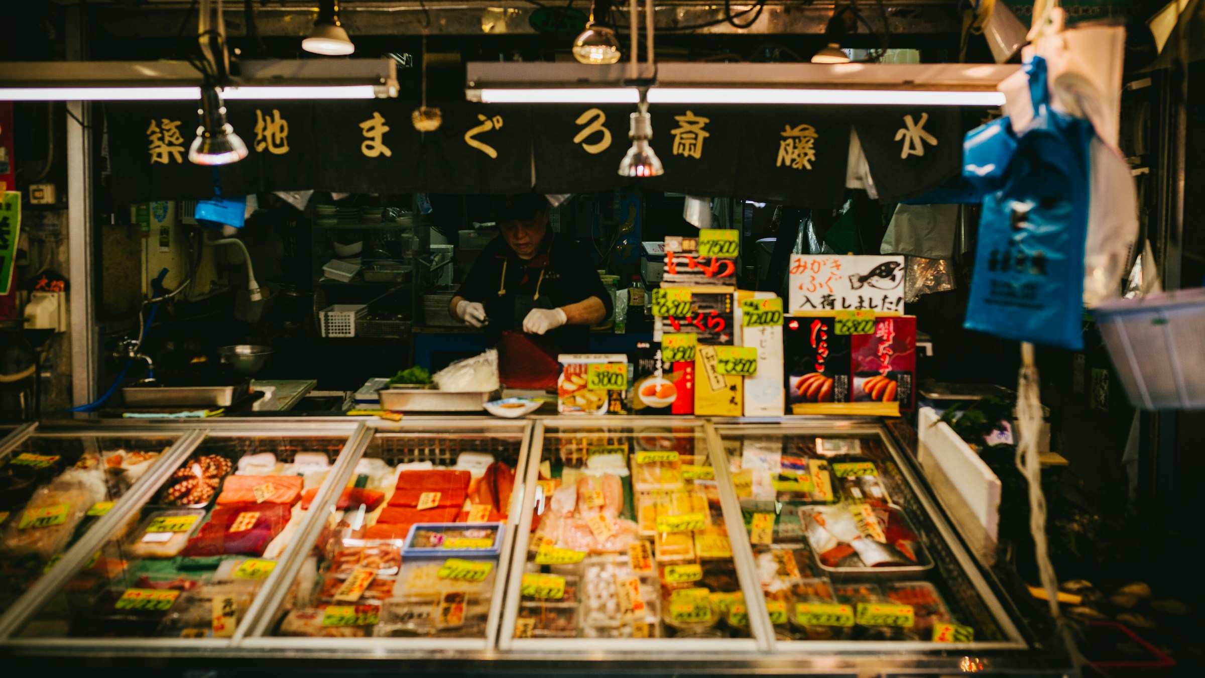 築地のまぐろ専門店「築地まぐろ斎藤水」の鮮魚陳列ケース。マグロ各部位のパックが並び、店主が奥に立つ。