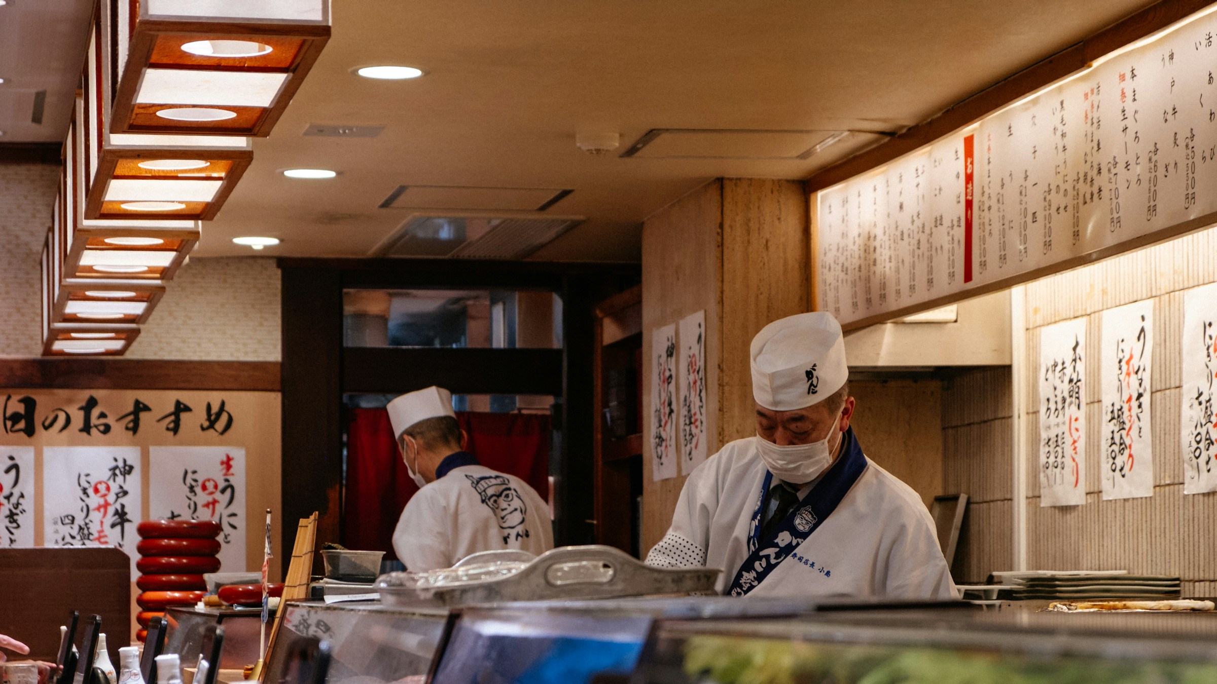 日本の街場の寿司店のカウンター。白衣と帽子の職人2名が握りを準備し、壁一面に達筆な日本語メニューが並ぶ。