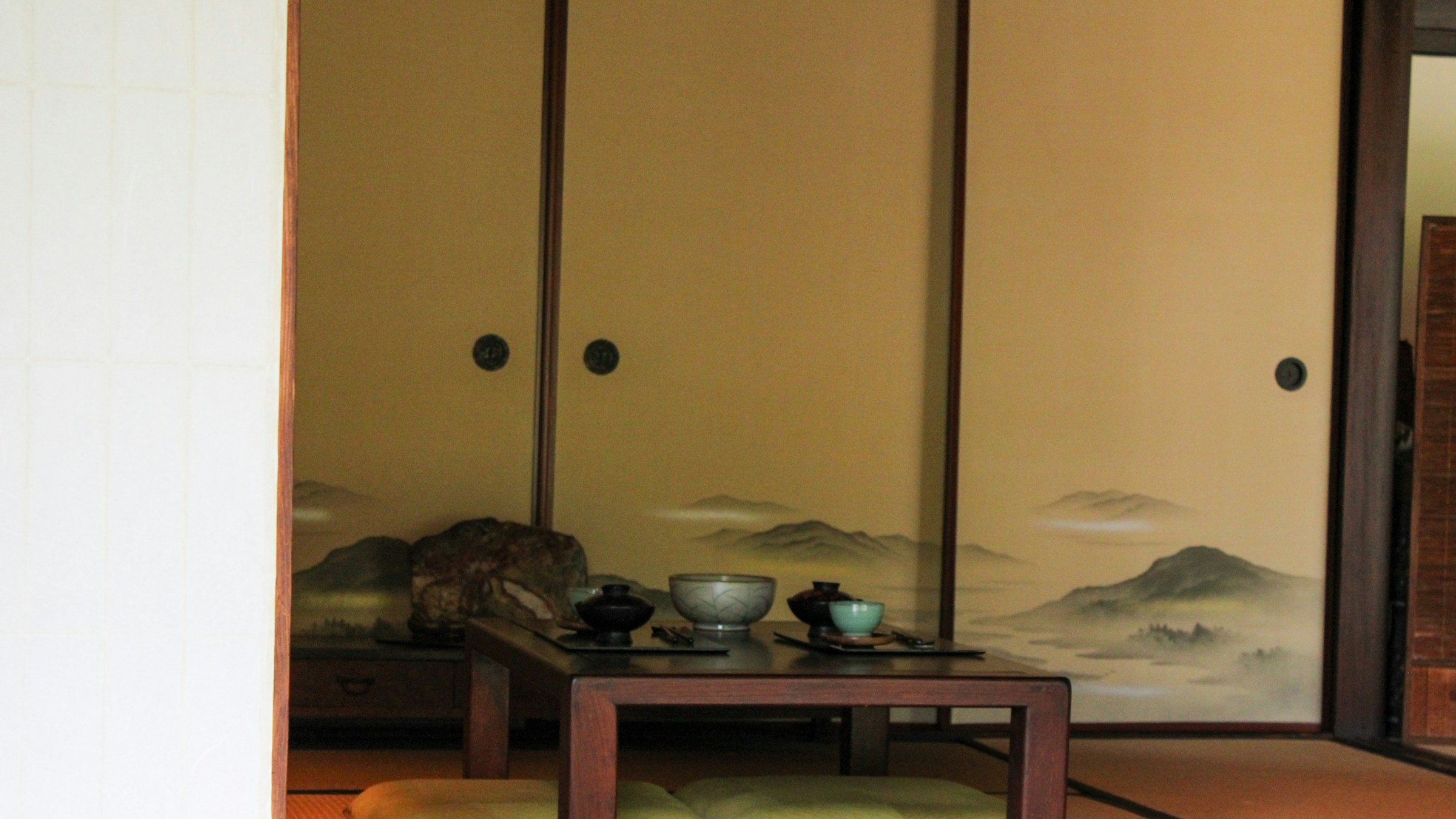 和室の床の間に山水画の襖、漆器の茶碗と抹茶椀が並ぶ茶席の設え。畳と座布団。