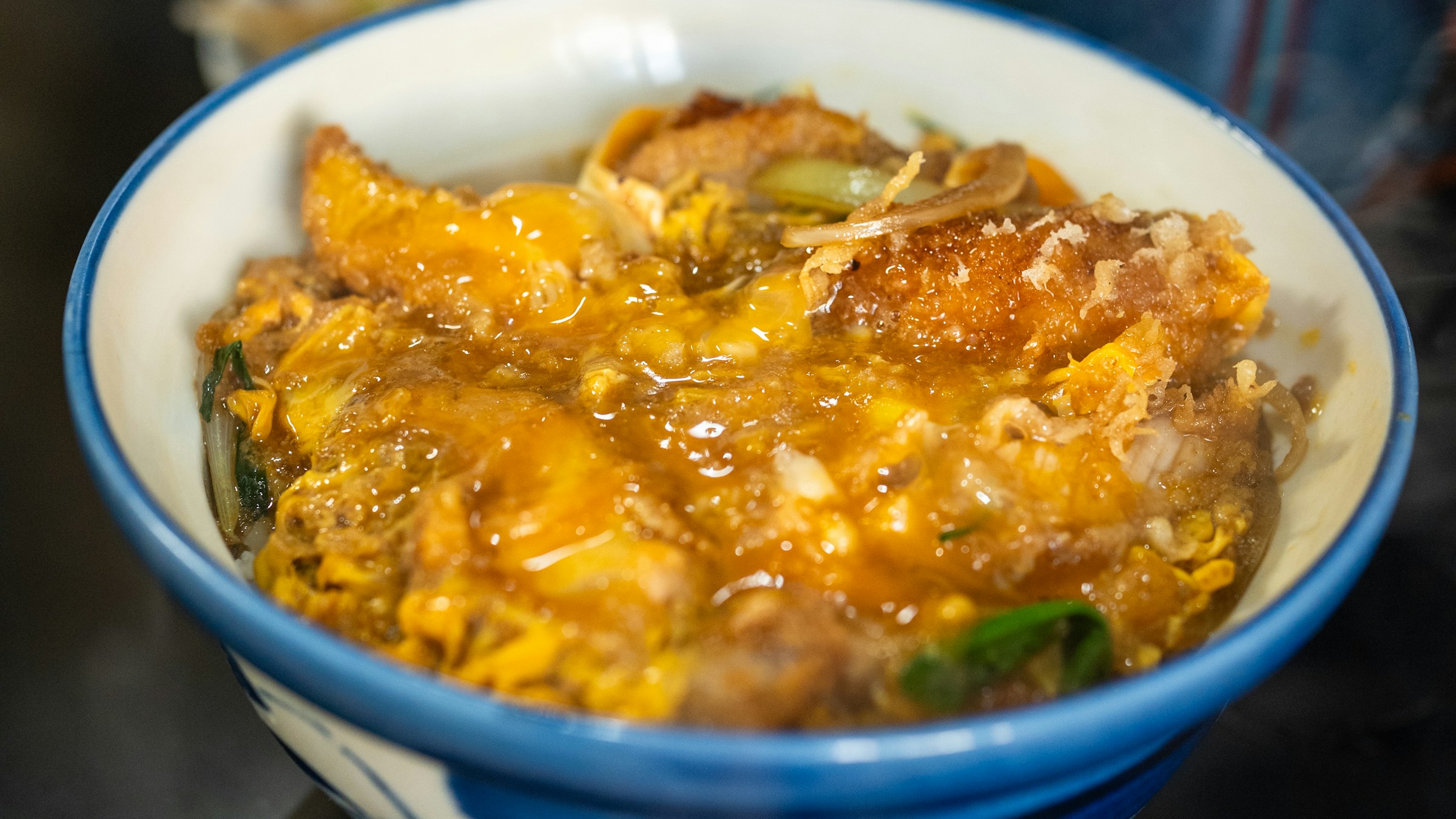 青と白の伝統的な日本の磁器丼に盛られた卵とじのカツ丼の上面クローズアップ。とんかつの上に卵と玉ねぎが絡み、緑のネギが散らされる。とんかつのカツ丼業態を象徴する一枚。