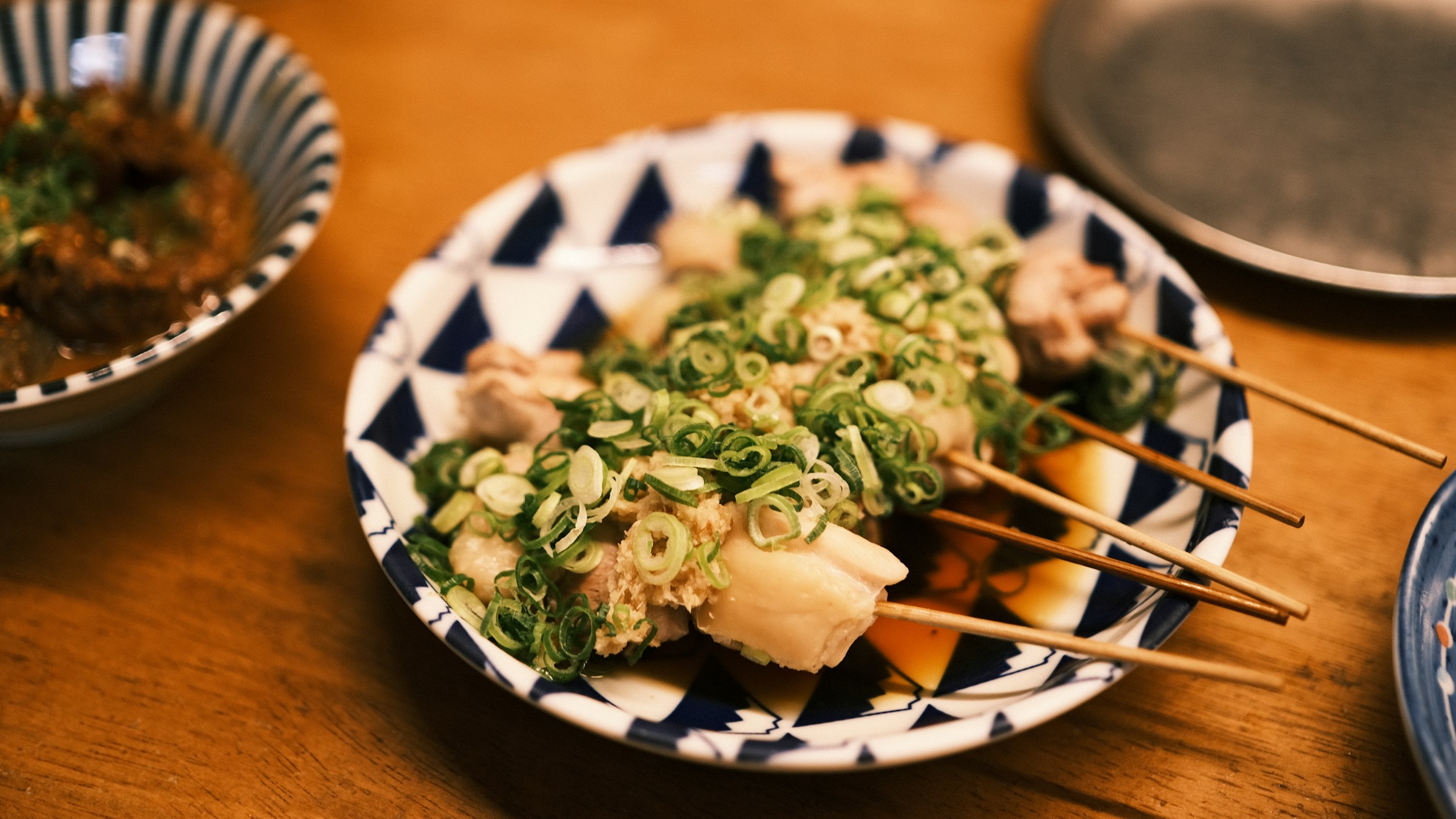 青と白の麻の葉柄の和食器に重ねて盛り付けられた焼鳥の串と、たっぷりの刻みネギ。木のテーブルの上、奥に薬味の小鉢。現代の焼鳥のねぎたっぷりスタイルを象徴する一枚。