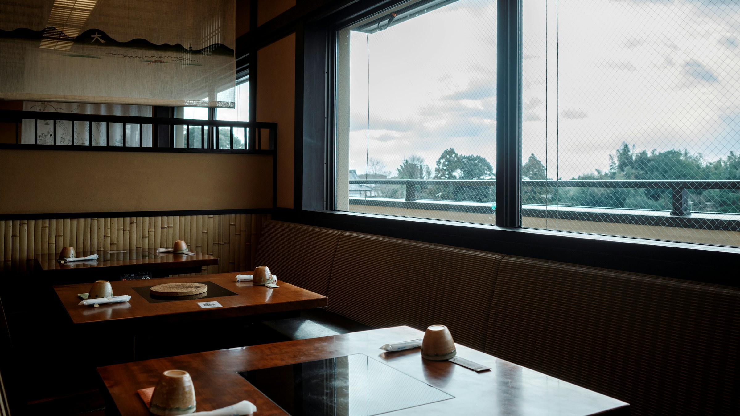 窓辺に川と樹々を望む和食店の店内。木のテーブルと竹のパーティション。