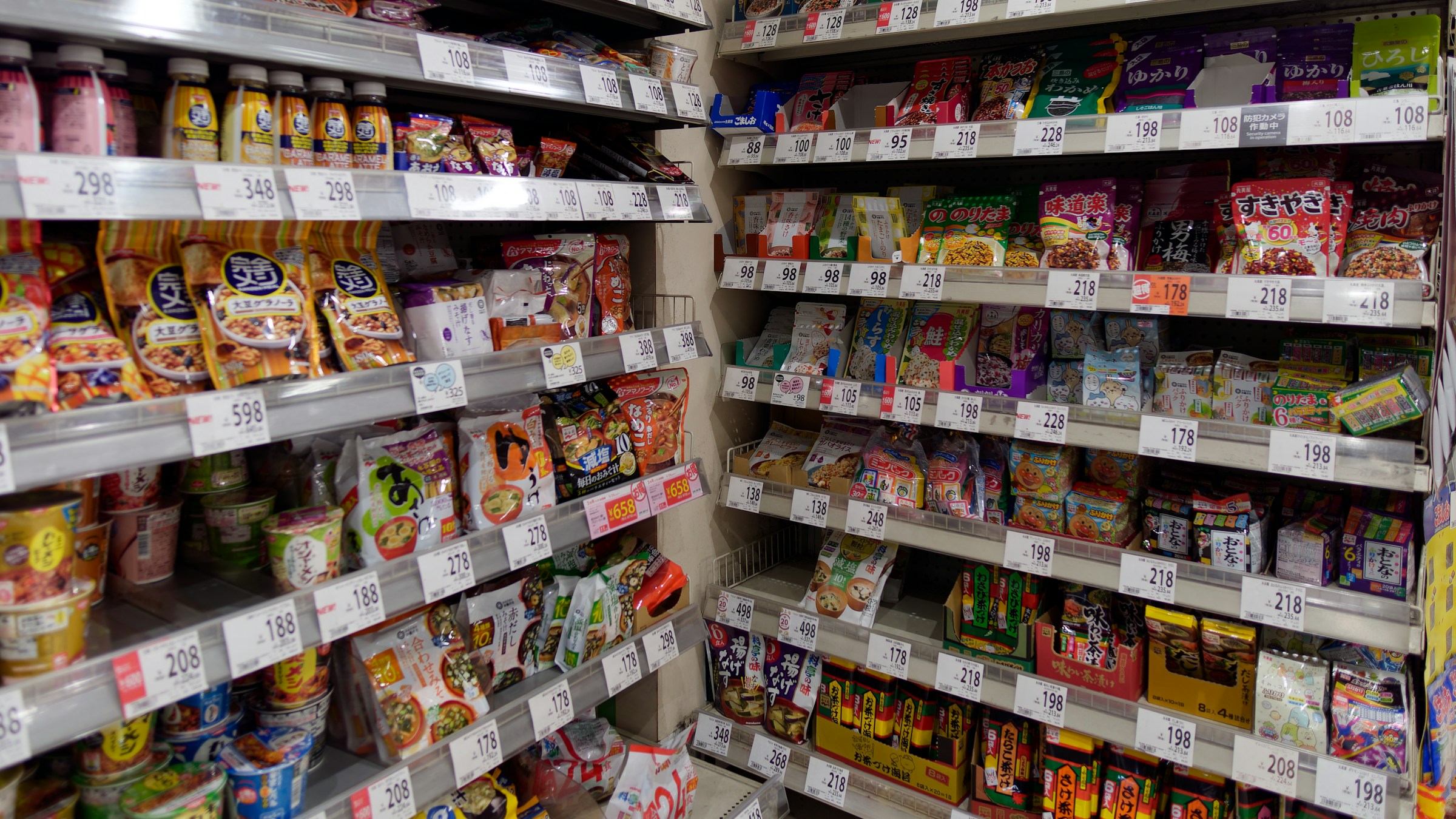 日本の店内の食品売場。カップ麺・スナック菓子・飲料が棚いっぱいに並び、価格(¥298、¥348、¥598、¥218 など)が黄色いPOPで表示されている。