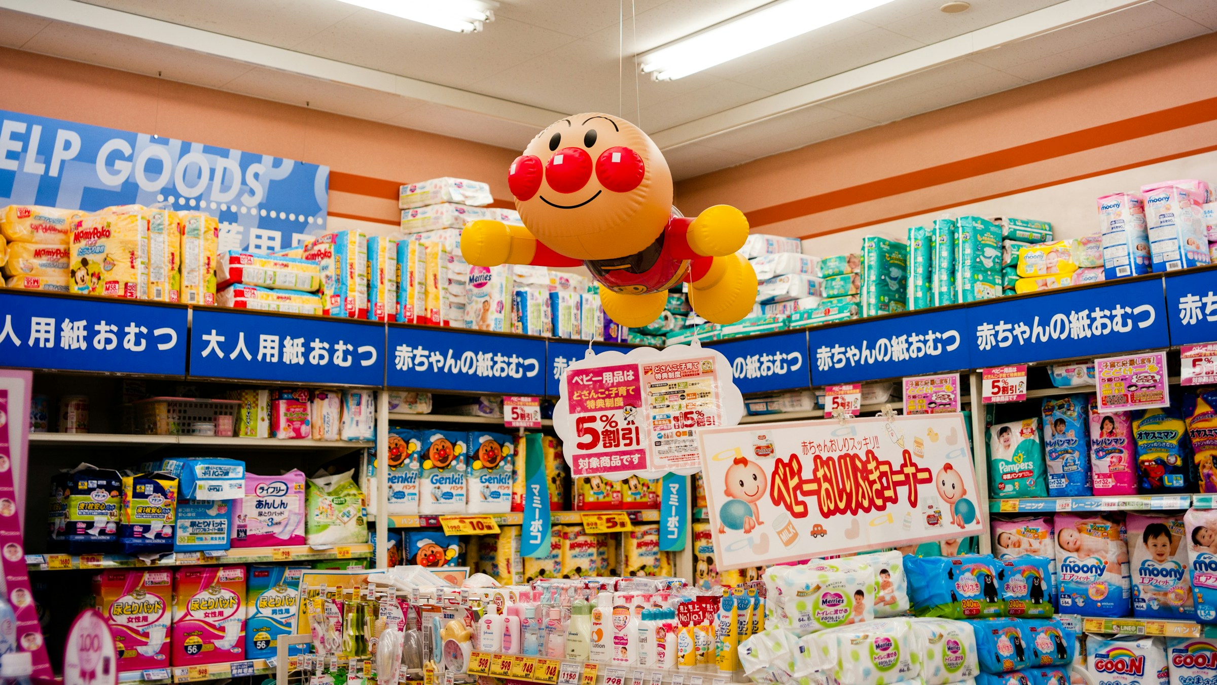 日本のドラッグストア店内。アンパンマンの風船が天井から下がり、棚にはパンパースやムーニーなどの紙おむつ、ベビー用品の特売告知。「ベビー紙おむつコーナー」「大人用紙おむつ」「赤ちゃんの紙おむつ」のPOPが並ぶ。